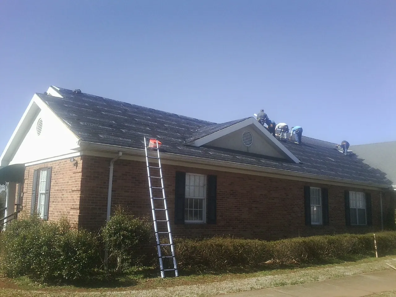 Crew tearing shingles off a brick ranch roof with a ladder set — Metal Roof Repair work by our crew in Nekoma