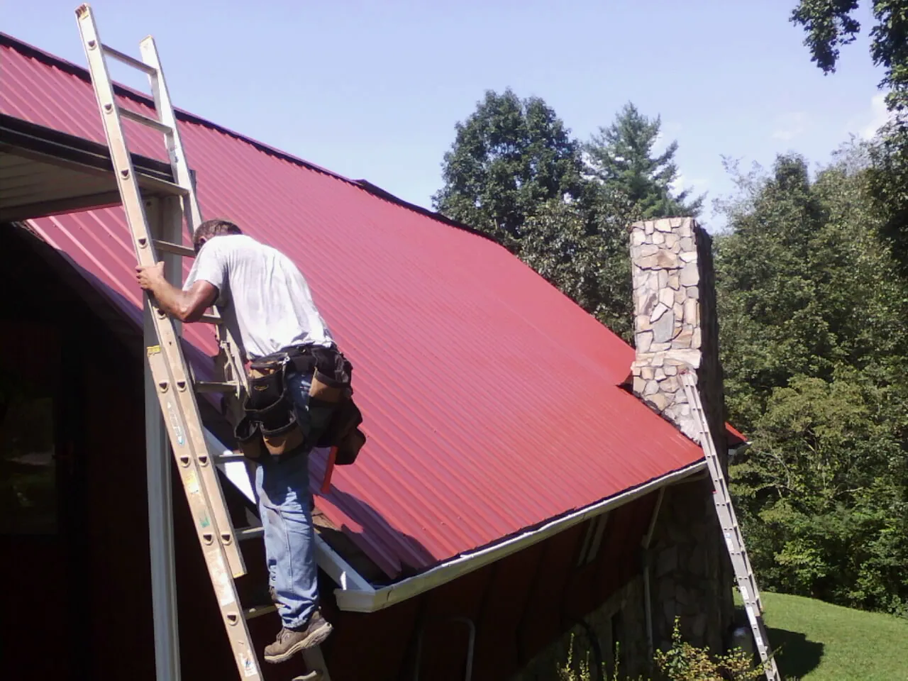 Roofer on a ladder beside a red metal roof and stone chimney — Soffit & Fascia Repair by Roofing Brothers in Nekoma
